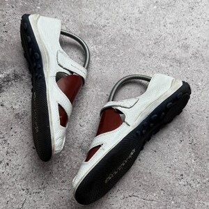 May include: A pair of white leather sandals featuring a dark brown interior, a black rubber outsole with a textured grip pattern, and a metallic silver shoe tree insert visible inside each shoe to maintain their shape.