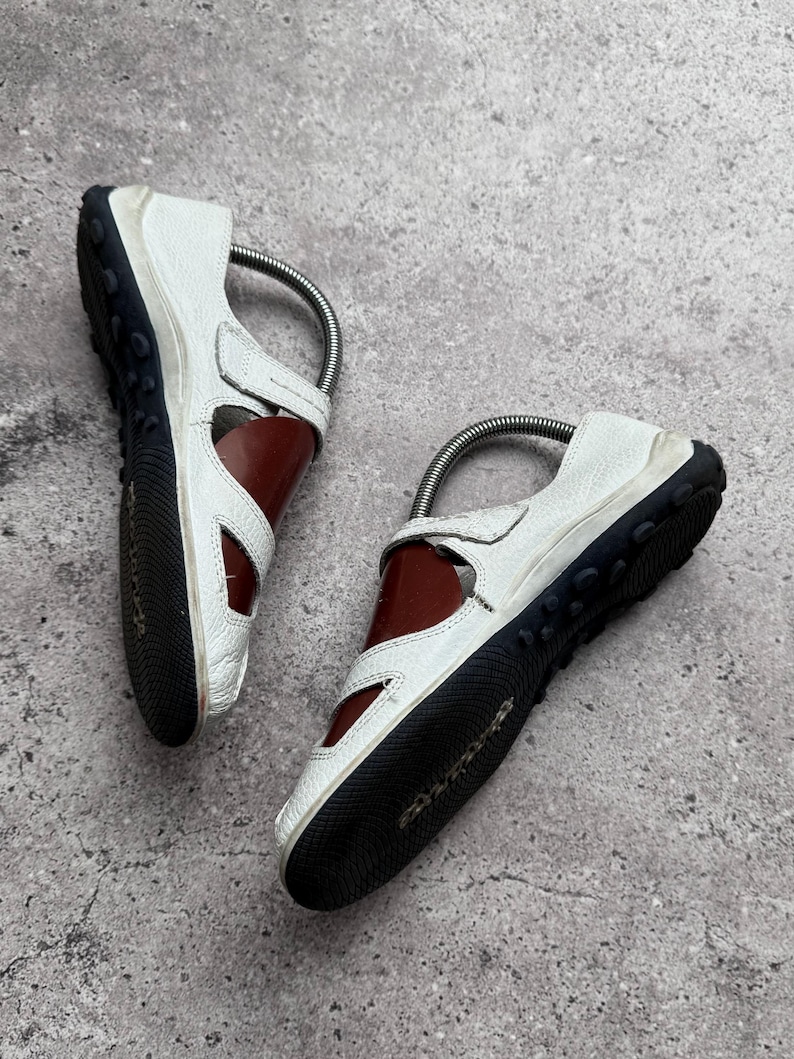 May include: A pair of white leather sandals featuring a dark brown interior, a black rubber outsole with a textured grip pattern, and a metallic silver shoe tree insert visible inside each shoe to maintain their shape.