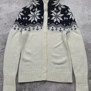 May include: A white and navy blue knit cardigan with a faux fur-lined hood. The upper portion features a snowflake pattern. The cardigan has a button-up front and ribbed cuffs. A cozy winter outerwear item.