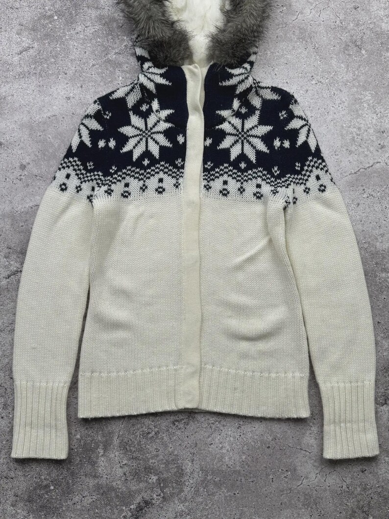 May include: A white and navy blue knit cardigan with a faux fur-lined hood. The upper portion features a snowflake pattern. The cardigan has a button-up front and ribbed cuffs. A cozy winter outerwear item.