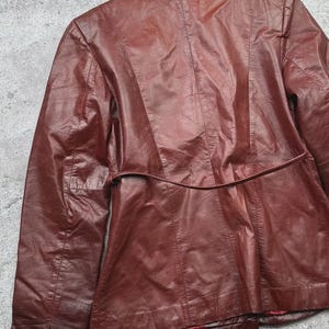 May include: A burgundy leather jacket, viewed from the back, showcasing a tailored design. The jacket features a notched collar, a belt detail at the waist, and vertical seams for a structured fit. The leather has a slightly worn appearance.