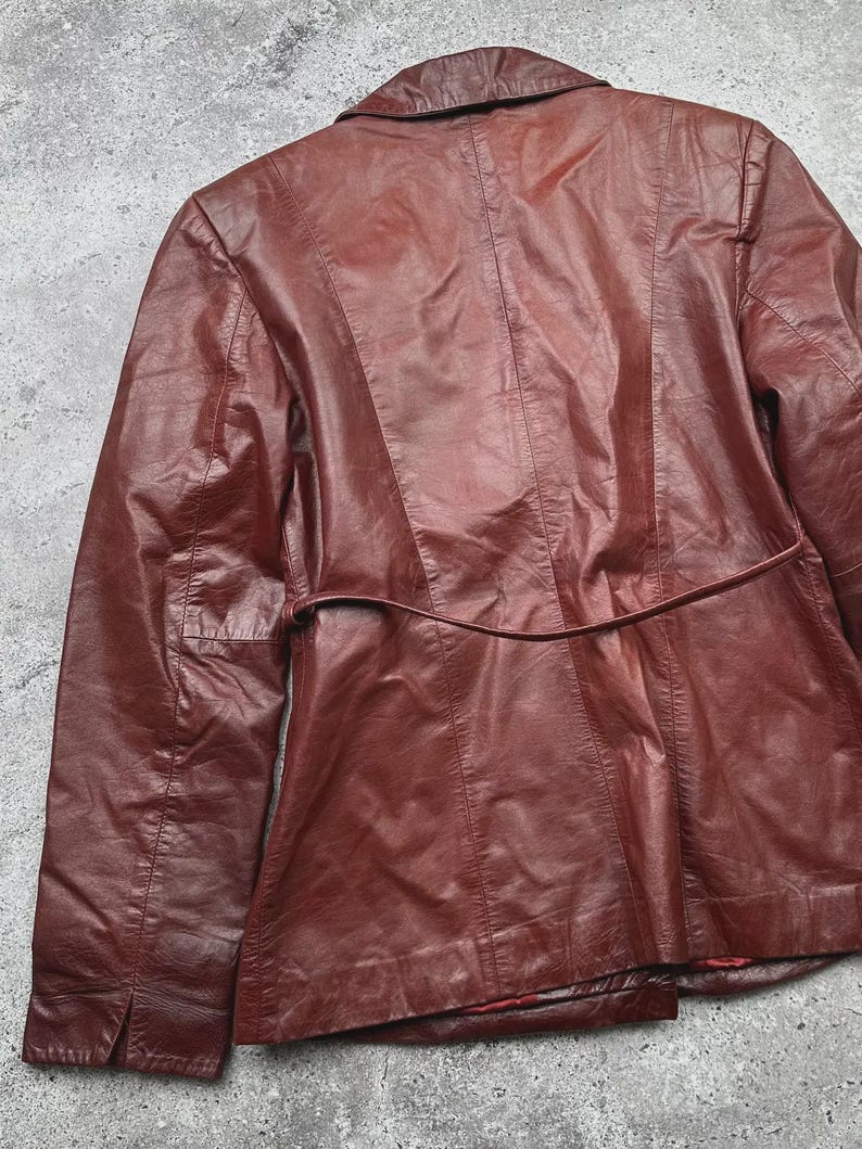 May include: A burgundy leather jacket, viewed from the back, showcasing a tailored design. The jacket features a notched collar, a belt detail at the waist, and vertical seams for a structured fit. The leather has a slightly worn appearance.