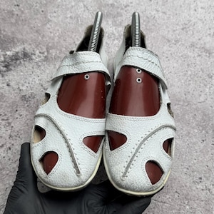 May include: A pair of white leather sandals featuring a closed-toe design with multiple cutouts, visible stitching along the edges, and dark brown shoe trees inserted inside to maintain the shape of the footwear.