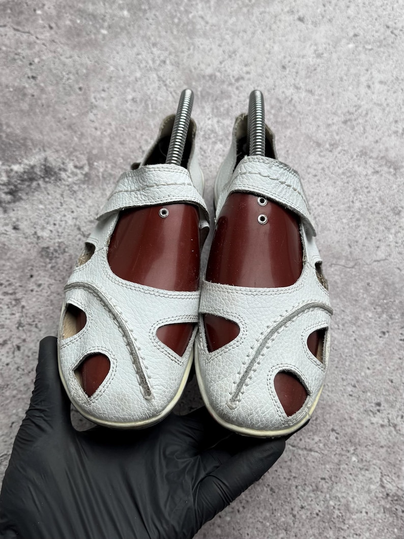 May include: A pair of white leather sandals featuring a closed-toe design with multiple cutouts, visible stitching along the edges, and dark brown shoe trees inserted inside to maintain the shape of the footwear.