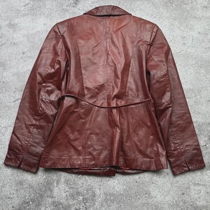 May include: A burgundy leather jacket with a classic design. The jacket features a notched collar, long sleeves, and a fitted waist with a belt detail. The back of the jacket has vertical seams for a tailored look. The leather has a slightly worn appearance.