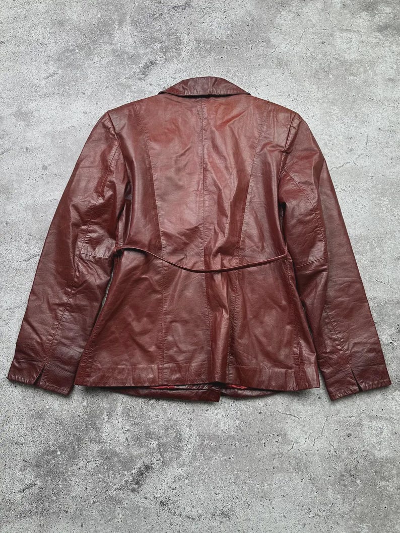 May include: A burgundy leather jacket with a classic design. The jacket features a notched collar, long sleeves, and a fitted waist with a belt detail. The back of the jacket has vertical seams for a tailored look. The leather has a slightly worn appearance.
