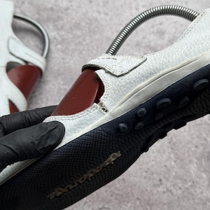 May include: A white textured leather shoe with a dark red side panel and a black rubber sole featuring a textured grip pattern and small circular studs. The shoe includes a metal spring shoe tree inserted inside to maintain its shape.