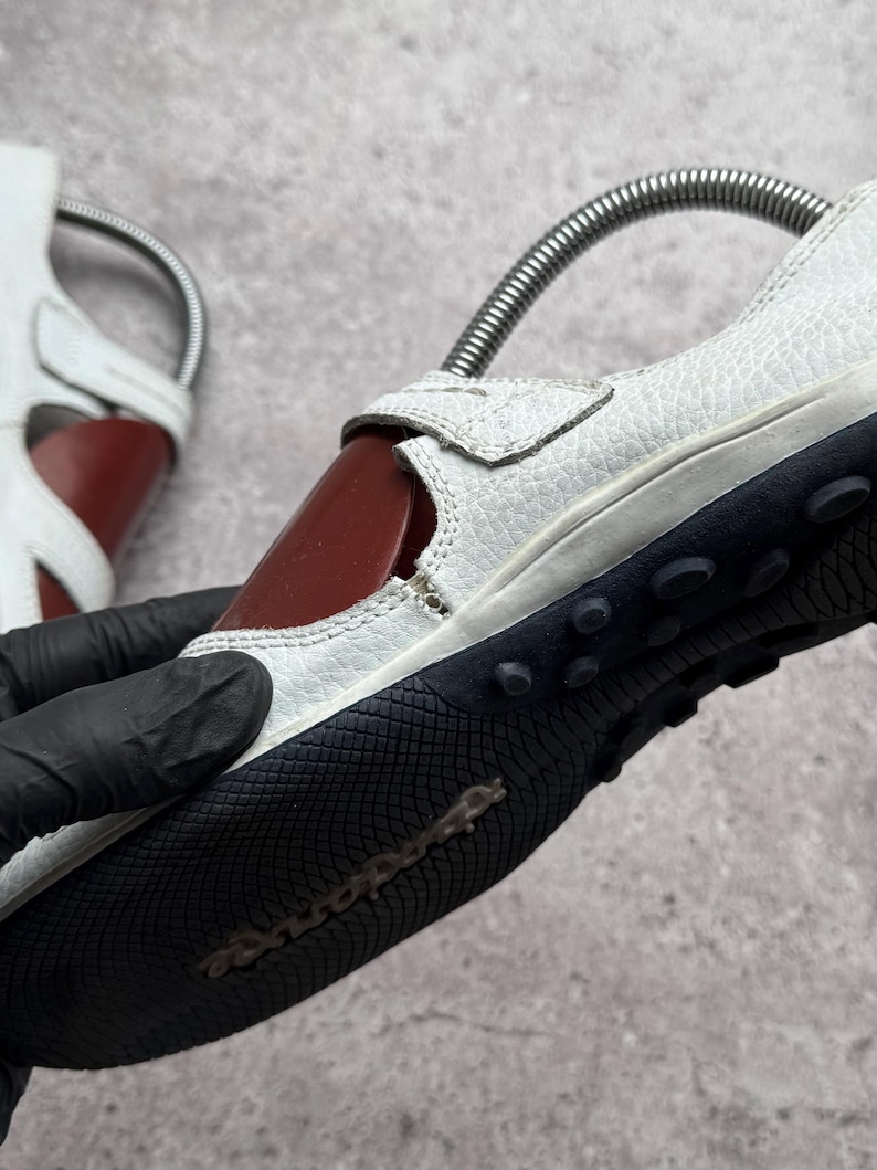 May include: A white textured leather shoe with a dark red side panel and a black rubber sole featuring a textured grip pattern and small circular studs. The shoe includes a metal spring shoe tree inserted inside to maintain its shape.