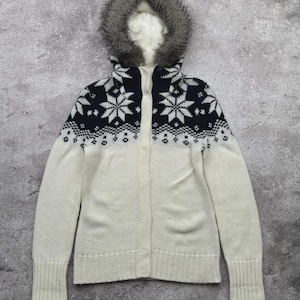 May include: A cream-colored zip-up sweater with a faux fur-lined hood. The upper portion features a navy blue snowflake pattern. The sweater has long sleeves and ribbed cuffs. A winter-themed garment.