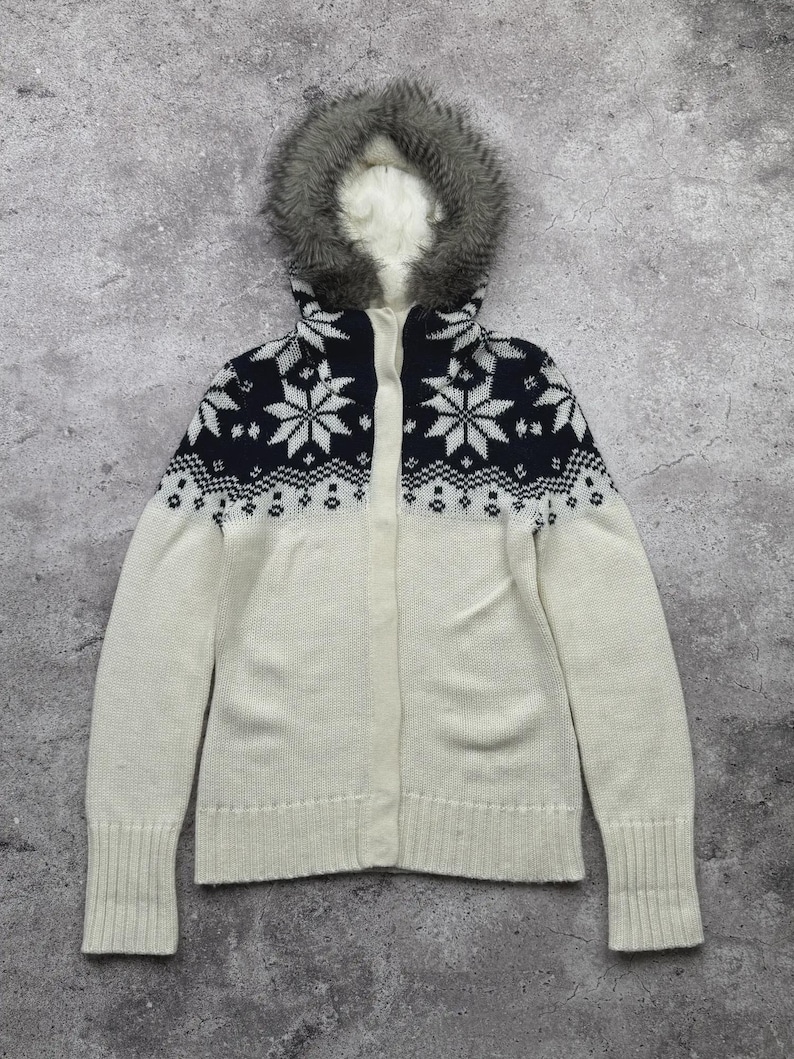May include: A cream-colored zip-up sweater with a faux fur-lined hood. The upper portion features a navy blue snowflake pattern. The sweater has long sleeves and ribbed cuffs. A winter-themed garment.