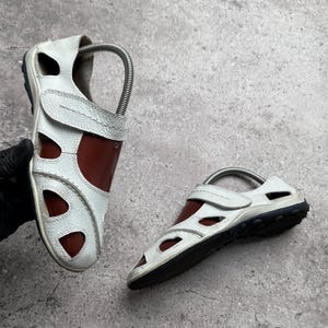 May include: A pair of white and brown closed-toe sandals featuring a textured white leather upper with cut-out panels, a brown inner lining, a hook-and-loop strap closure, and a black rubber sole with a textured tread pattern for traction.