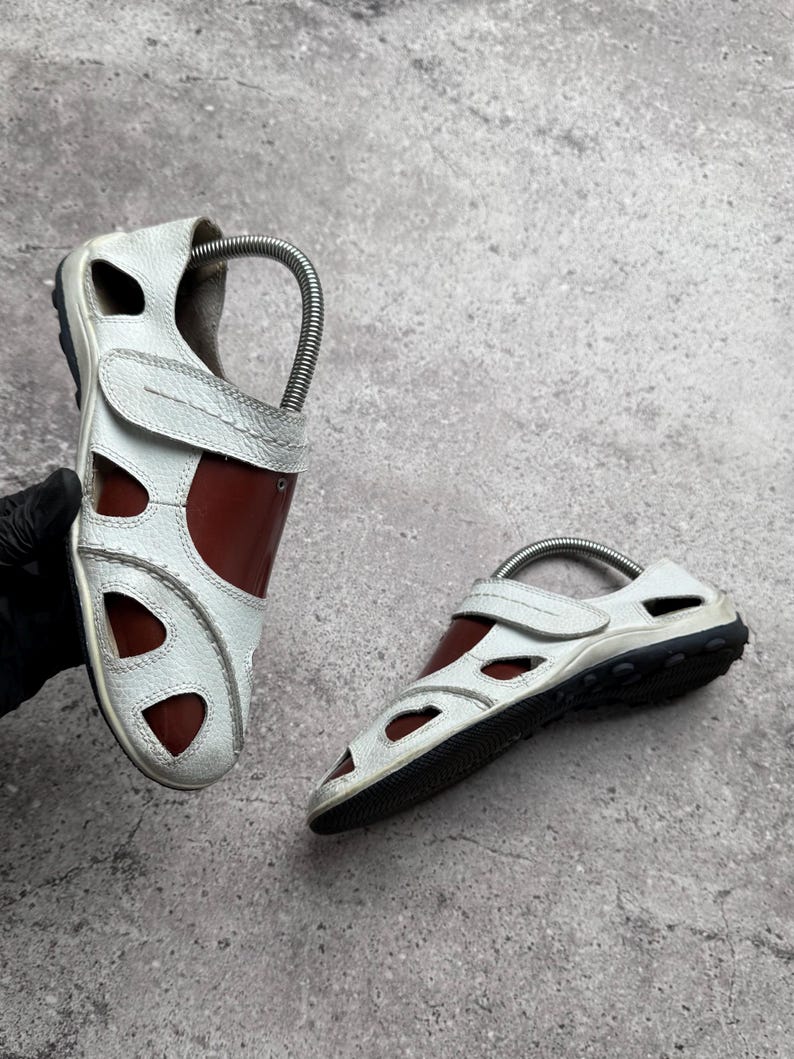 May include: A pair of white and brown closed-toe sandals featuring a textured white leather upper with cut-out panels, a brown inner lining, a hook-and-loop strap closure, and a black rubber sole with a textured tread pattern for traction.