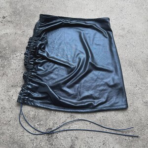 May include: A black, faux leather mini skirt with a ruched side detail. The skirt has a slight sheen and a drawstring closure. The fabric appears to be smooth and drapes softly. The skirt is a solid color and has a simple, modern design.