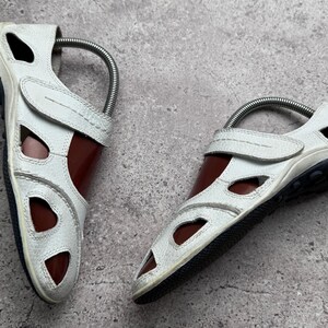 May include: A pair of white leather sandals featuring a closed-toe design with multiple cutouts, a hook-and-loop strap closure across the top, and a dark brown interior lining. The sandals have a textured white exterior and a black rubber sole.