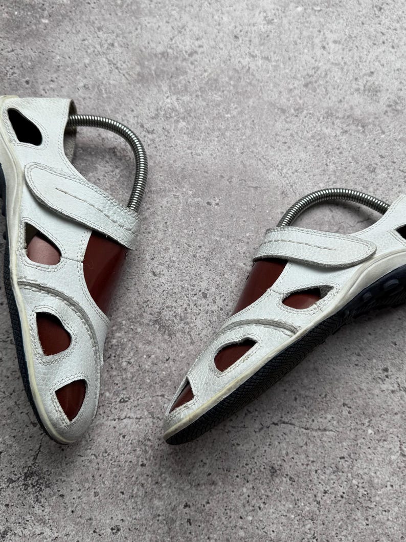 May include: A pair of white leather sandals featuring a closed-toe design with multiple cutouts, a hook-and-loop strap closure across the top, and a dark brown interior lining. The sandals have a textured white exterior and a black rubber sole.
