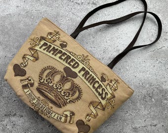 Pode incluir: Uma bolsa tote de lona bege com alças de couro marrom. A bolsa apresenta uma coroa marrom com uma faixa que diz "Pampered Princess" e o texto "I shop therefore I am".