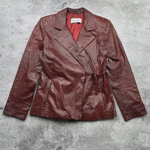 May include: A burgundy leather jacket with a wrap-style closure and a tie belt. The jacket features a notched collar and a red lining. The sleeves are long, and the jacket appears to be vintage.