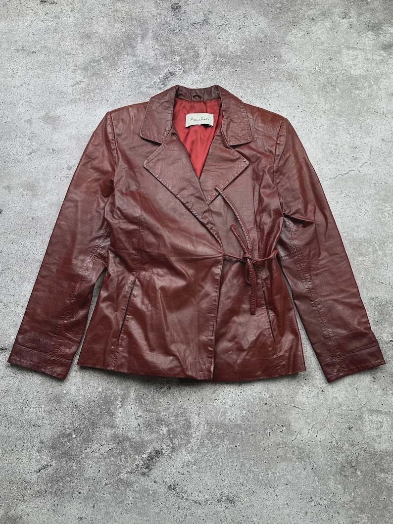 May include: A burgundy leather jacket with a wrap-style closure and a tie belt. The jacket features a notched collar and a red lining. The sleeves are long, and the jacket appears to be vintage.