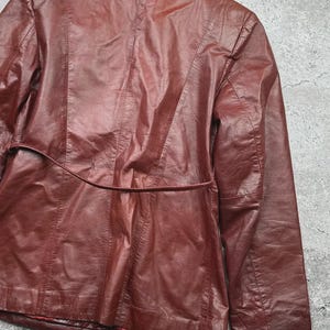 May include: A burgundy leather jacket with a tailored fit. The jacket features a notched collar, long sleeves, and a belt detail at the waist. The back of the jacket has vertical seams and a split at the bottom.