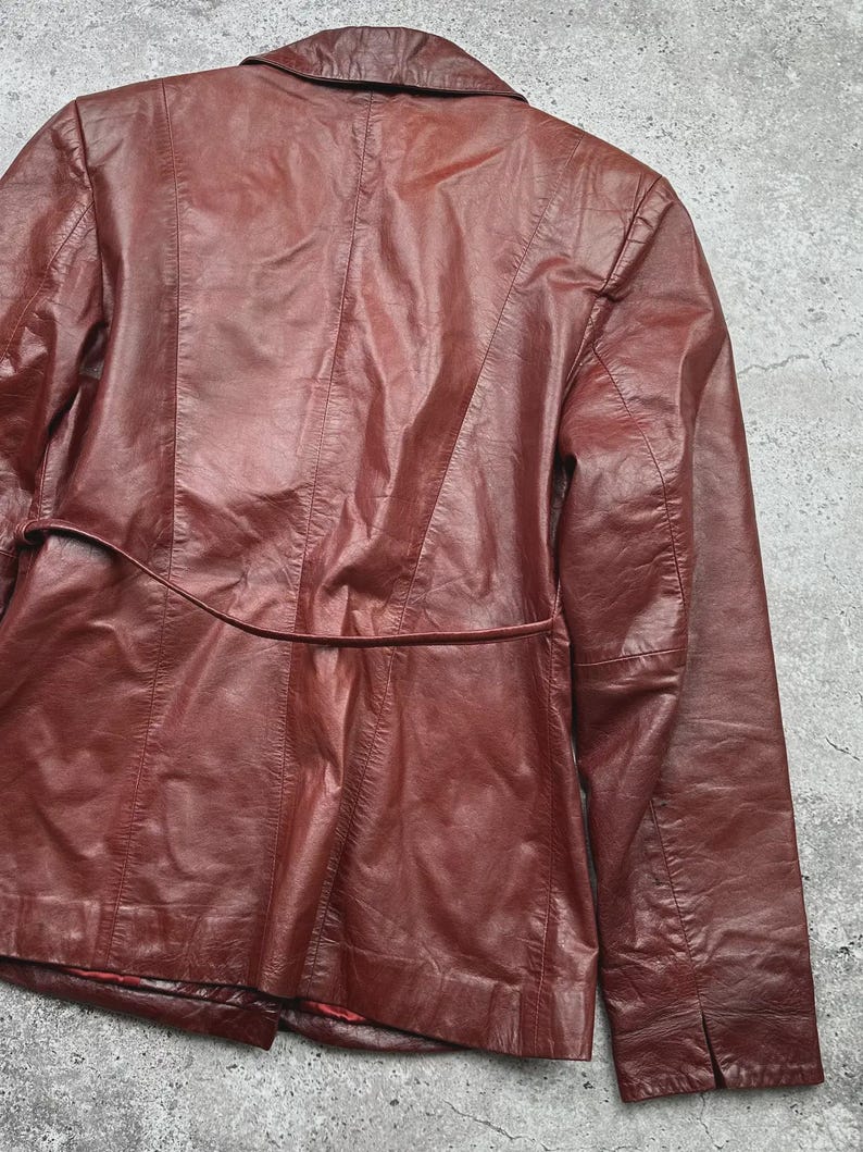 May include: A burgundy leather jacket with a tailored fit. The jacket features a notched collar, long sleeves, and a belt detail at the waist. The back of the jacket has vertical seams and a split at the bottom.