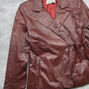 May include: A burgundy leather jacket with a notched collar and a tie closure at the waist. The jacket has long sleeves and a red satin lining. The label inside the jacket reads "Pellino".