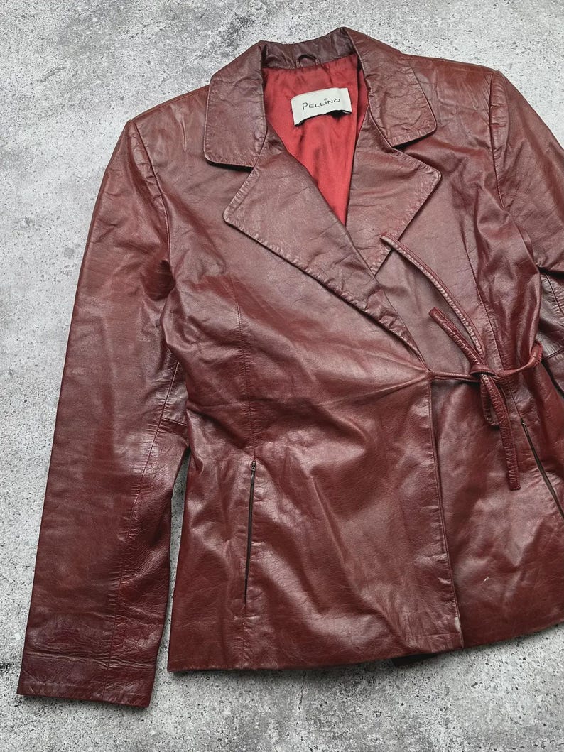 May include: A burgundy leather jacket with a notched collar and a tie closure at the waist. The jacket has long sleeves and a red satin lining. The label inside the jacket reads "Pellino".