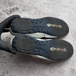 May include: A pair of white shoes with black rubber outsoles featuring a textured tread pattern and a central logo. The soles have a rounded heel and toe design with a series of small, raised circular grips along the perimeter of the black base.
