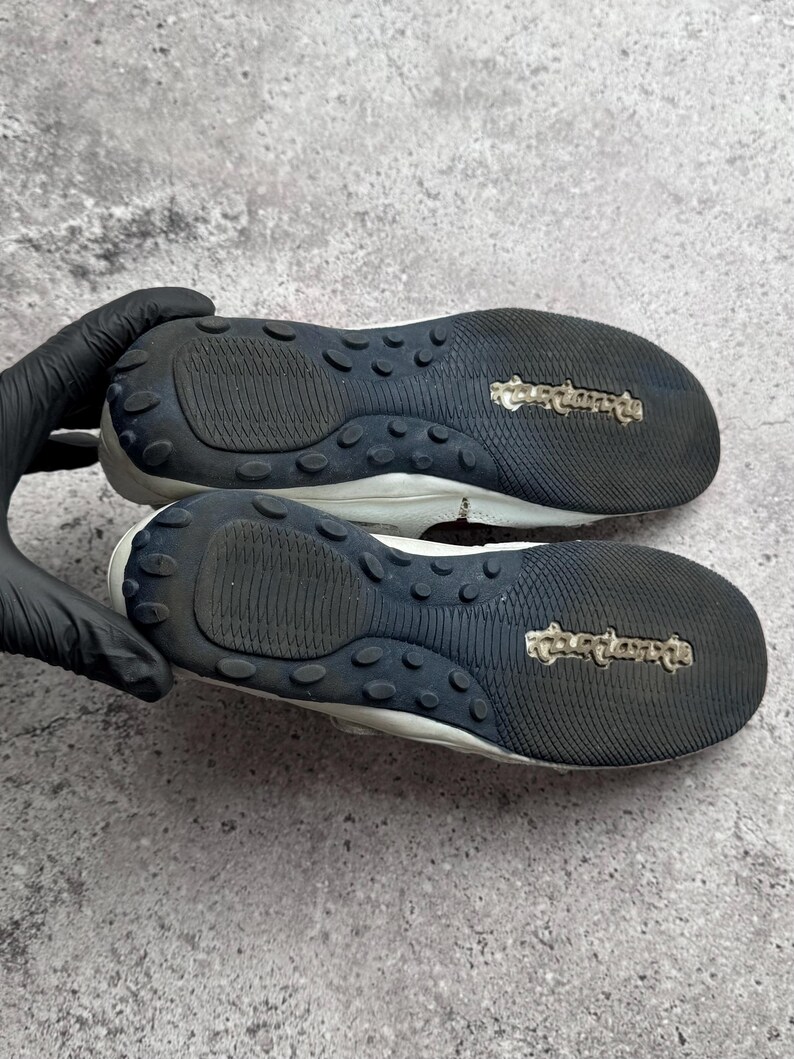 May include: A pair of white shoes with black rubber outsoles featuring a textured tread pattern and a central logo. The soles have a rounded heel and toe design with a series of small, raised circular grips along the perimeter of the black base.
