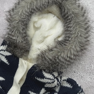 May include: A close-up of a winter coat hood with a faux fur trim in shades of gray and white. The hood's interior is lined with soft, white fabric. The coat features a navy blue and white snowflake pattern.