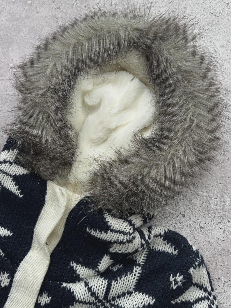 May include: A close-up of a winter coat hood with a faux fur trim in shades of gray and white. The hood's interior is lined with soft, white fabric. The coat features a navy blue and white snowflake pattern.