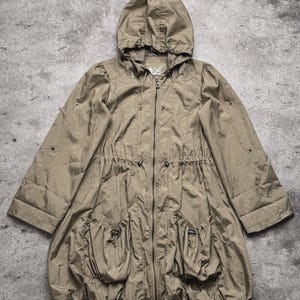 May include: A taupe hooded jacket featuring a full-length front zipper, a drawstring waist, and two large front pockets with drawstring closures. The jacket has long sleeves with adjustable tabs and a gathered, rounded hemline design.