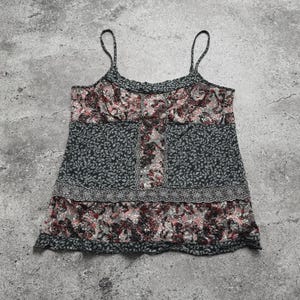 May include: A floral print camisole top with thin spaghetti straps. The top features a mix of dark gray and floral patterns in shades of pink, red, and white. The top has a ruffled neckline and a decorative border.