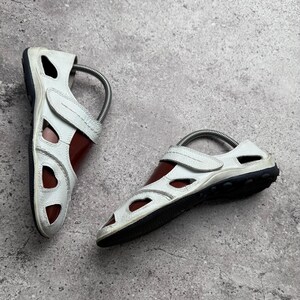 May include: A pair of white leather sandals featuring a closed-toe design with multiple cutouts, a hook-and-loop strap closure across the instep, and a dark brown sole. Each sandal includes a metal spring shoe tree inserted for shape retention.
