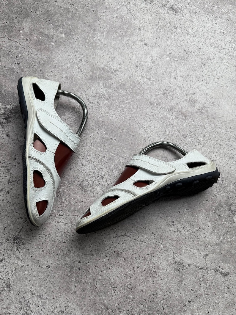 May include: A pair of white leather sandals featuring a closed-toe design with multiple cutouts, a hook-and-loop strap closure across the instep, and a dark brown sole. Each sandal includes a metal spring shoe tree inserted for shape retention.
