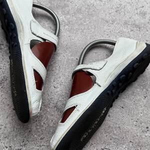 May include: A pair of white leather shoes with cut-out side panels, featuring a dark brown interior lining and a black rubber sole with a textured tread pattern. Each shoe includes a metal shoe tree insert to maintain its shape and structure.