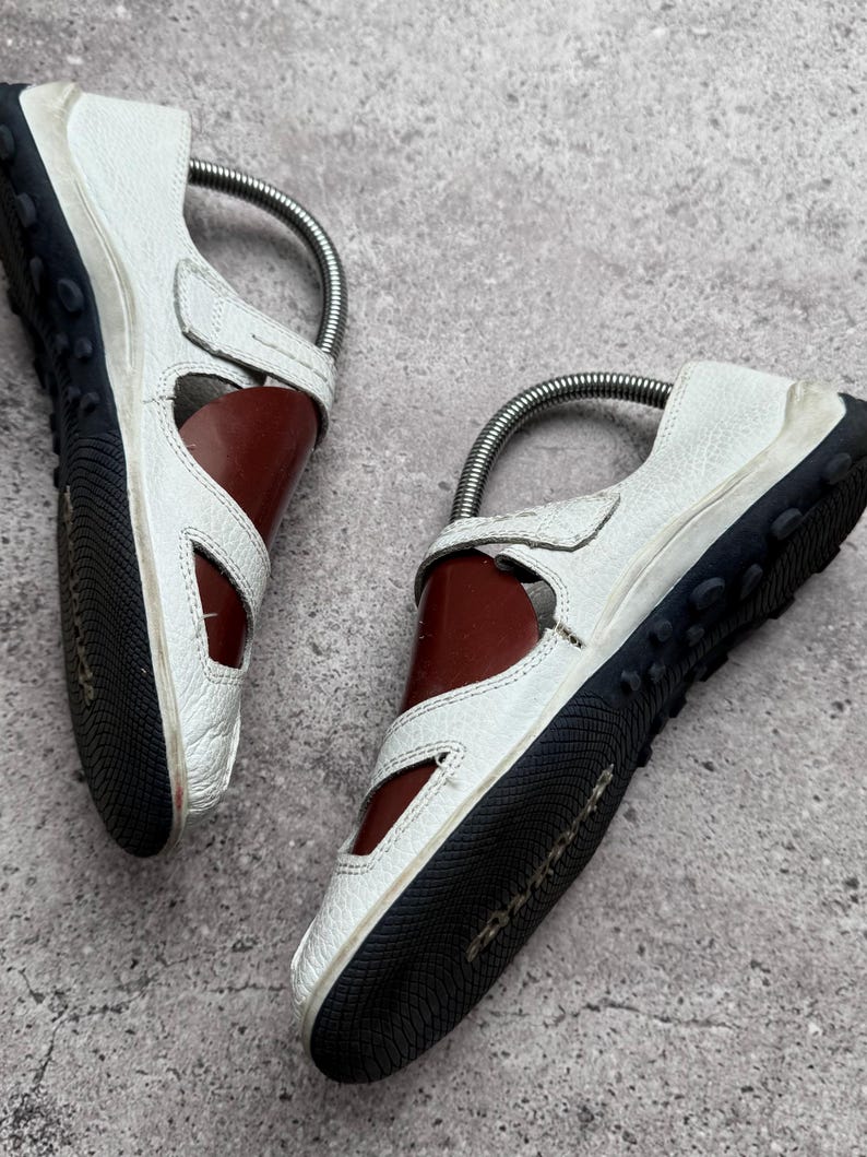 May include: A pair of white leather shoes with cut-out side panels, featuring a dark brown interior lining and a black rubber sole with a textured tread pattern. Each shoe includes a metal shoe tree insert to maintain its shape and structure.