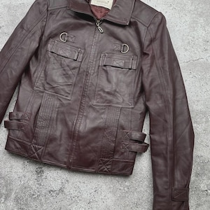K&ouml;nnte beinhalten: Burgunderfarbene Lederjacke mit Rei&szlig;verschluss, zwei Brusttaschen und zwei unteren Taschen. Die Jacke hat einen Kragen, D-Ring-Details und verstellbare Riemen an Taille und Manschetten. Die Jacke ist aus glattem, geschmeidigem Leder gefertigt.