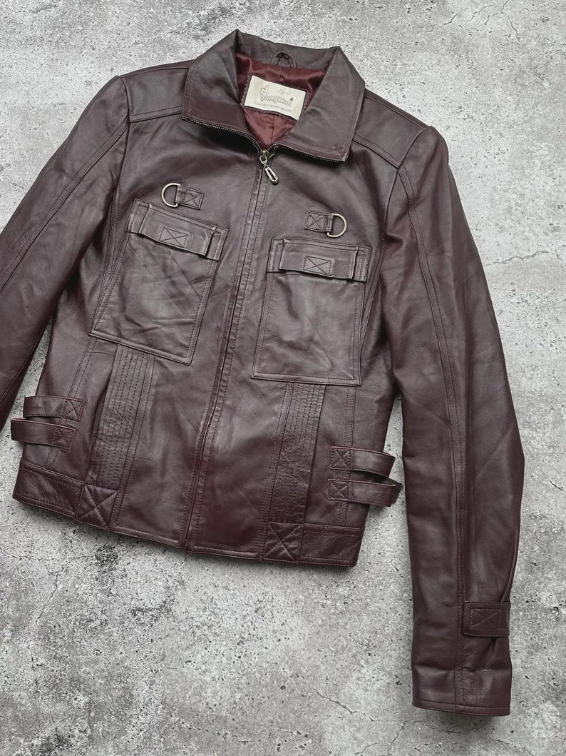 K&ouml;nnte beinhalten: Burgunderfarbene Lederjacke mit Rei&szlig;verschluss, zwei Brusttaschen und zwei unteren Taschen. Die Jacke hat einen Kragen, D-Ring-Details und verstellbare Riemen an Taille und Manschetten. Die Jacke ist aus glattem, geschmeidigem Leder gefertigt.