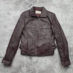 K&ouml;nnte beinhalten: Dunkelbraune Lederjacke mit Rei&szlig;verschluss, zwei Brusttaschen und zwei unteren Taschen. Die Jacke hat einen Kragen und dekorative Schnallen an Taille und Manschetten. Mittlere Gr&ouml;&szlig;e.