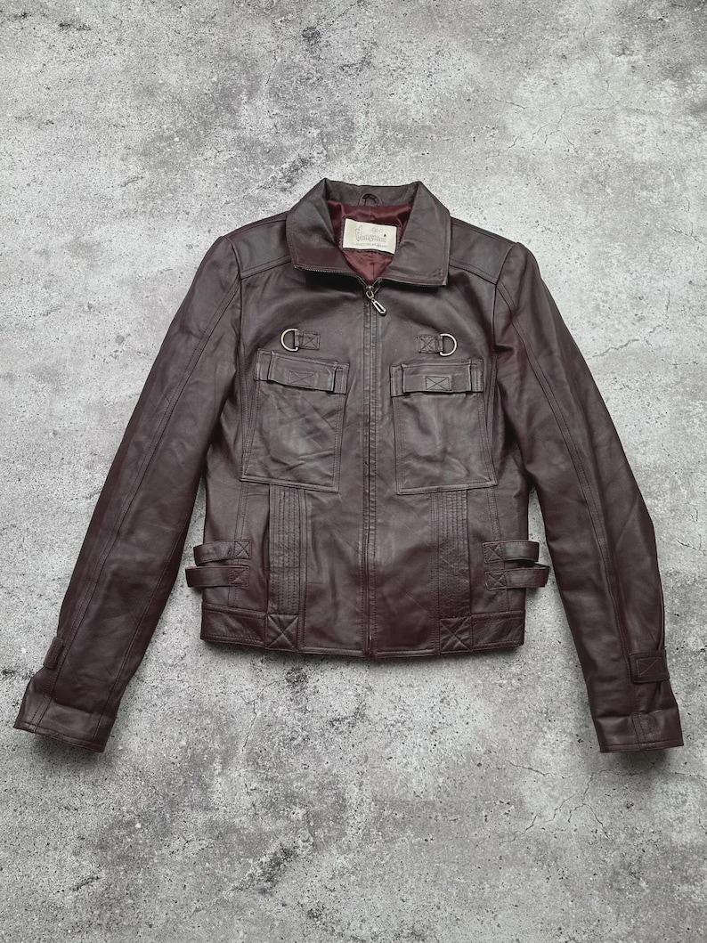 K&ouml;nnte beinhalten: Dunkelbraune Lederjacke mit Rei&szlig;verschluss, zwei Brusttaschen und zwei unteren Taschen. Die Jacke hat einen Kragen und dekorative Schnallen an Taille und Manschetten. Mittlere Gr&ouml;&szlig;e.