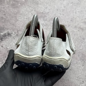 May include: A pair of white textured leather sandals featuring a closed heel design, a hook-and-loop strap closure, and a dark navy blue rubber outsole with a prominent circular tread pattern, shown from the back with metal shoe trees inside.