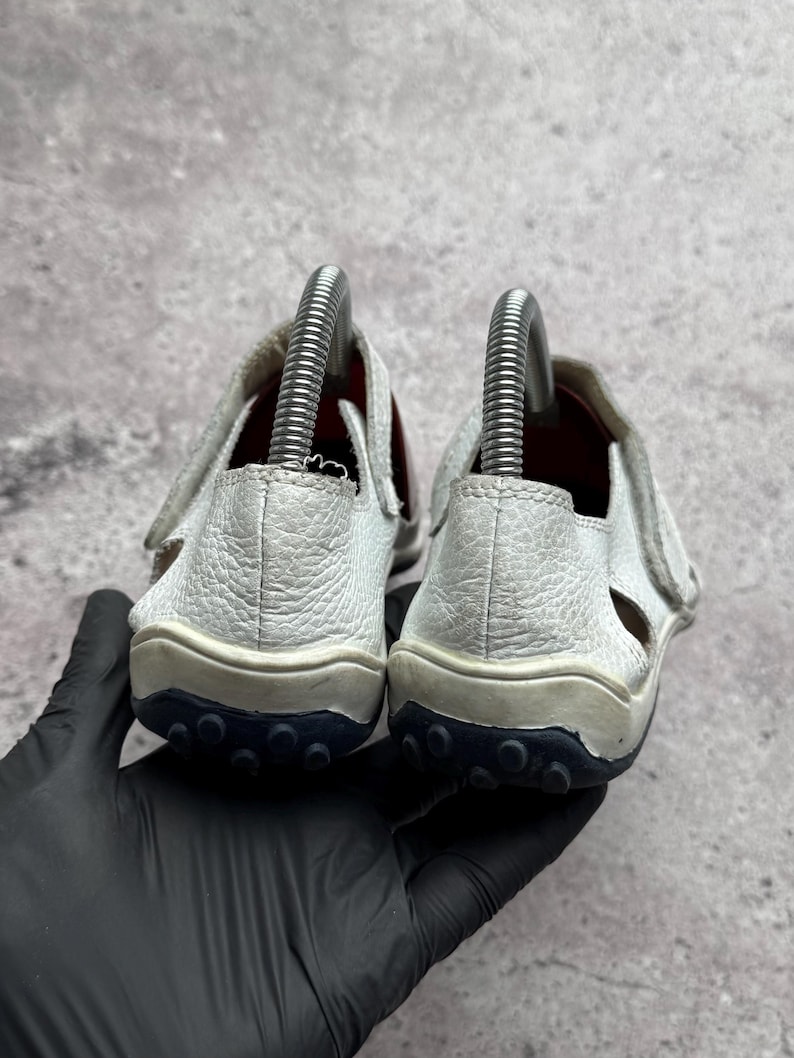 May include: A pair of white textured leather sandals featuring a closed heel design, a hook-and-loop strap closure, and a dark navy blue rubber outsole with a prominent circular tread pattern, shown from the back with metal shoe trees inside.