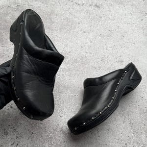 May include: A pair of black leather clogs featuring a rounded toe, a slip-on design, and a thick sole. The upper part of the shoe is accented with a row of small silver-toned metal studs along the base, providing a distinct decorative detail.