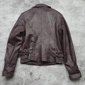K&ouml;nnte beinhalten: Braune Lederjacke mit klassischem Design. Die Jacke hat einen Kragen, einen Rei&szlig;verschluss und dekorative Schnallen an Taille und &Auml;rmeln. Die R&uuml;ckansicht zeigt die N&auml;hte und die Struktur der Jacke.