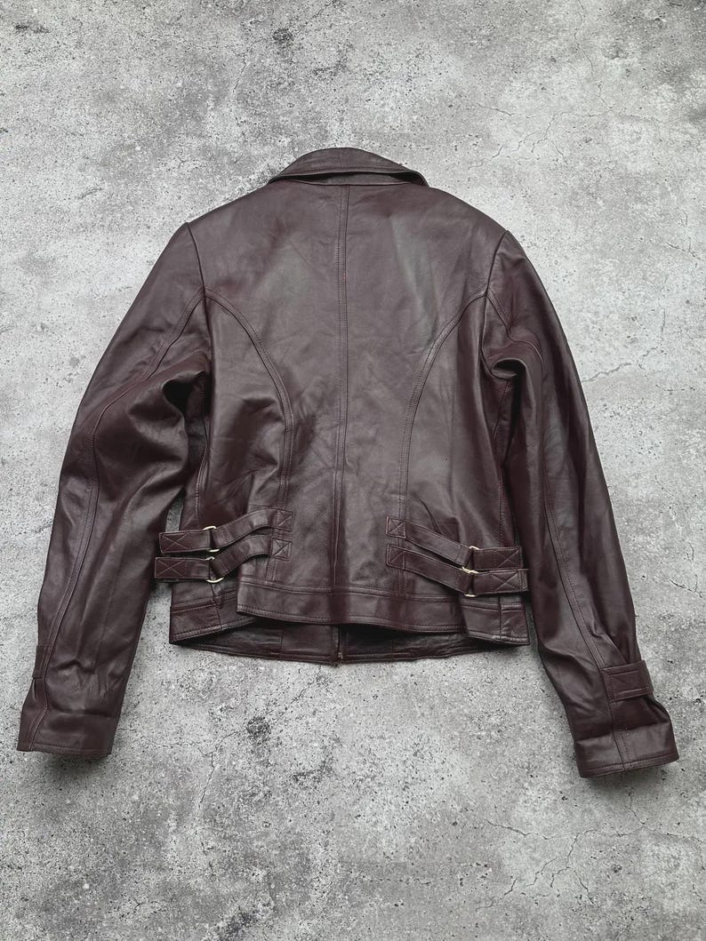 K&ouml;nnte beinhalten: Braune Lederjacke mit klassischem Design. Die Jacke hat einen Kragen, einen Rei&szlig;verschluss und dekorative Schnallen an Taille und &Auml;rmeln. Die R&uuml;ckansicht zeigt die N&auml;hte und die Struktur der Jacke.