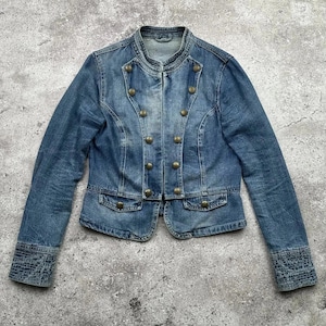 Könnte beinhalten: Blaue Jeansjacke mit Stehkragen und messingfarbenen Knöpfen. Die Jacke hat ein doppelreihiges Design, zwei kleine Taschen und dekorative Nähte an den Manschetten. Die Jacke ist aus mittelblauem Denim.