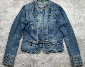 May include: A blue denim jacket with a mandarin collar and brass-colored buttons. The jacket has a double-breasted design, two small pockets, and decorative stitching on the cuffs. The jacket is a medium wash denim.
