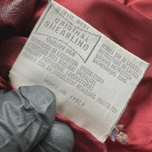 May include: Close-up of a tag from a garment. The tag reads "THE BEST ORIGINAL SHEARLING" and "MADE IN ITALY". The tag is white with black text and is attached to a burgundy-colored fabric.