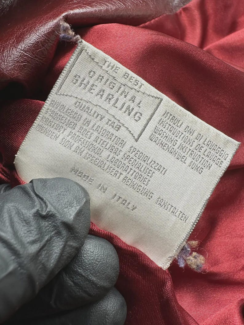May include: Close-up of a tag from a garment. The tag reads "THE BEST ORIGINAL SHEARLING" and "MADE IN ITALY". The tag is white with black text and is attached to a burgundy-colored fabric.