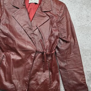 May include: A burgundy leather jacket with a tie belt closure. The jacket has a notched collar and a red satin lining. The brand name "Pellino" is visible on a tag inside the jacket.